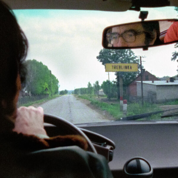Claude Lanzmann au volant d’une voiture à l’entrée du village de Treblinka en face de la gare. Treblinka, Pologne, juillet 1978.

Ce panneau a bouleversé Claude Lanzmann. Dans ses Mémoires, il écrit : « Je repris la route, continuant à conduire très lentement, et soudain j'aperçus une pancarte avec des lettres noires sur fond jaune qui indiquaient, comme si de rien n'était, le nom du village dans lequel nous entrions : «TREBLINKA». Autant j'étais resté insensible devant la douce pente enneigée du camp, ses stèles et son blockhaus central qui prétendait marquer l'emplacement des chambres à gaz, autant ce simple panneau d'ordinaire signalisation routière me mit en émoi. Treblinka existait ! Un village nommé Treblinka existait. Osait exister. Cela me semblait impossible. »